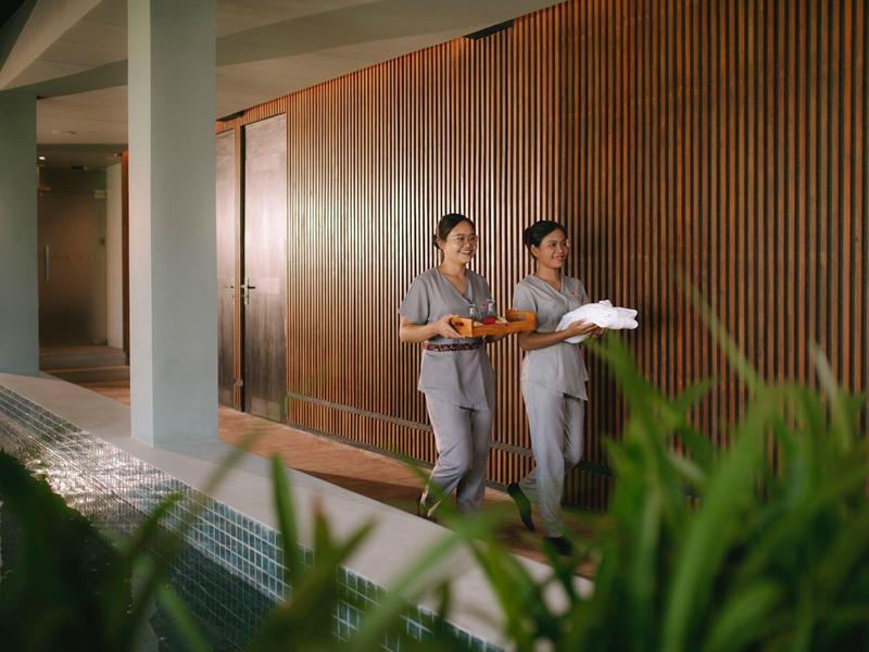 Two hotel staff carrying trays with drinks along a water channel and wooden paneling.