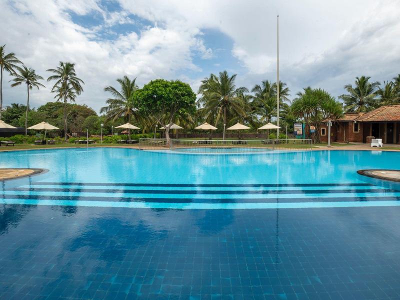 Large hotel pool with palm trees, sun umbrellas, and blue sky in the background.