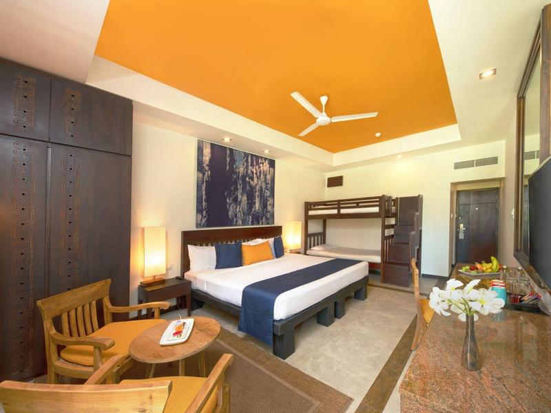 Modern hotel room with double bed, wooden chairs, large window, and orange ceiling accent.