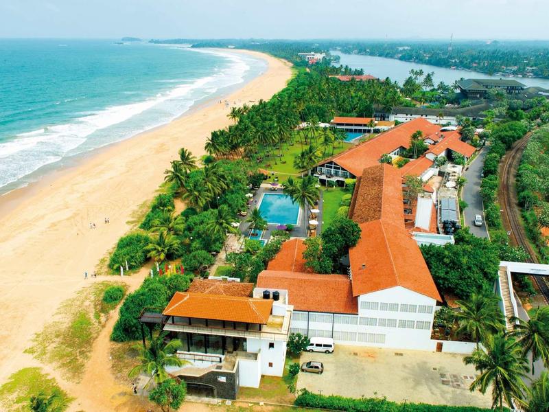 Luchtfoto van een kustresort met rode daken aan het strand met palmbomen langs de kust.