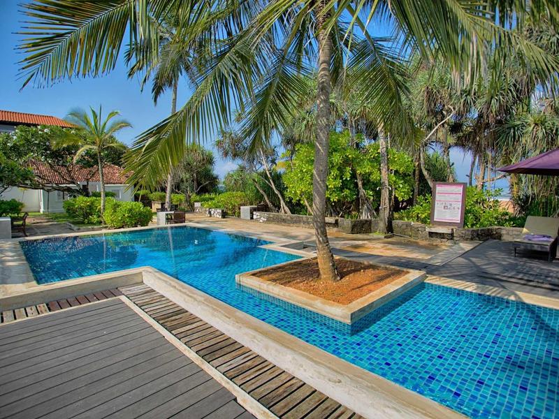 Hotelzwembad met blauwe tegels en palmbomen, schaduwrijke zitplaatsen en tropische omgeving.