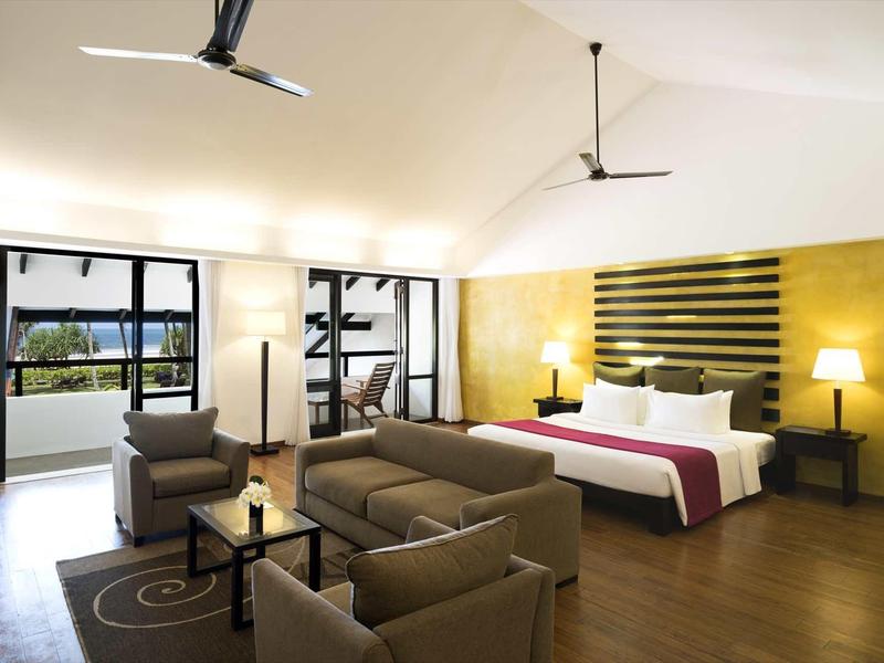 Ruime hotelkamer met tweepersoonsbed, bank, fauteuils en grote ramen met uitzicht naar buiten.