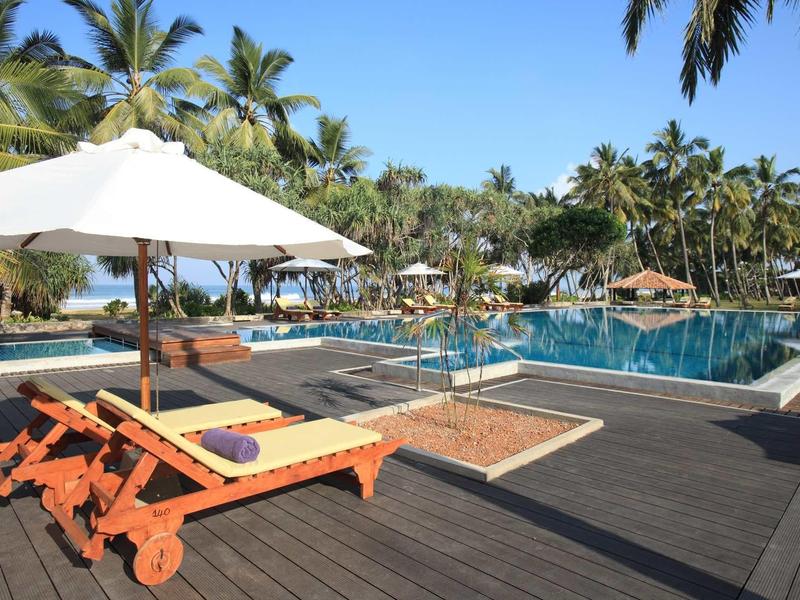 Zonterras met ligstoelen en zwembad, omgeven door palmbomen onder een heldere hemel.