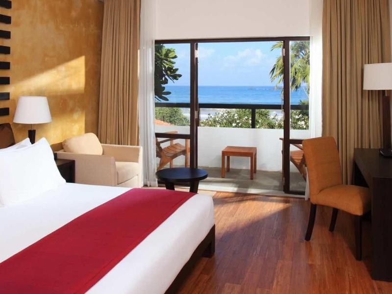 Moderne hotelkamer met tweepersoonsbed, fauteuils en zeezicht door grote ramen.