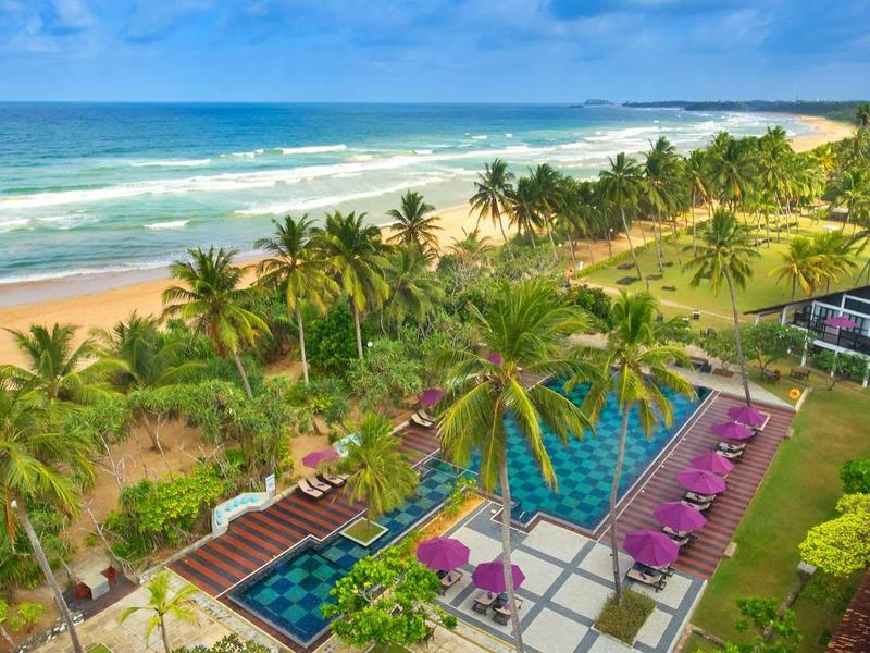 Uitzicht op een tropisch strand met palmbomen, zwembaden en ligstoelen bij heldere lucht.