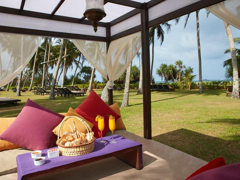 Gezellige strandlounge met kussens, fruit en uitzicht op palmbomen en oceaan.