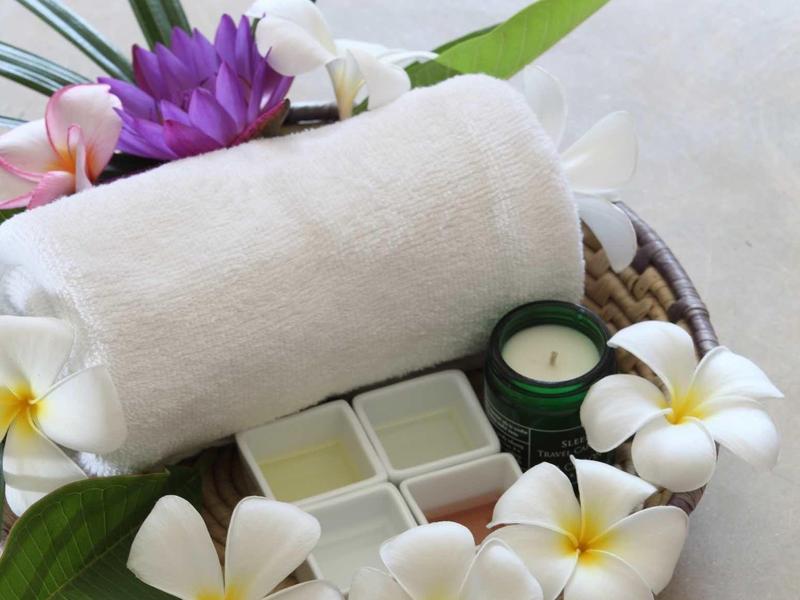 Wellnessmand met witte handdoek, kaars en witte bloemen op een lichte ondergrond.