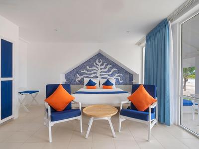 Wohnzimmer mit blauen Sesseln, orangen Kissen, rundem Holztisch und blauer Wanddekoration.