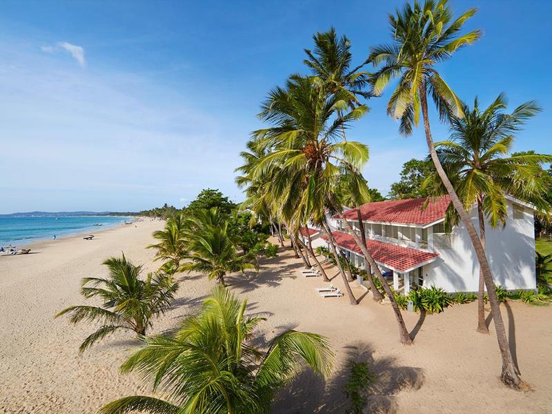 Ein weißes Haus mit rotem Dach steht neben Palmen am Sandstrand unter blauem Himmel.