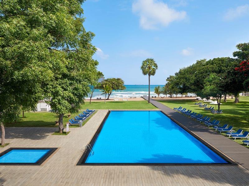 Großer rechteckiger Pool mit blauem Wasser, umgeben von Liegestühlen und grünen Bäumen am Meer.