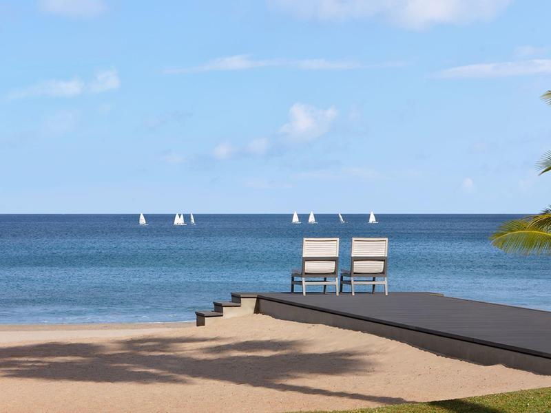 Zwei Stühle am Holzsteg am Strand, ruhiges Meer mit Segelbooten und blauem Himmel im Hintergrund.