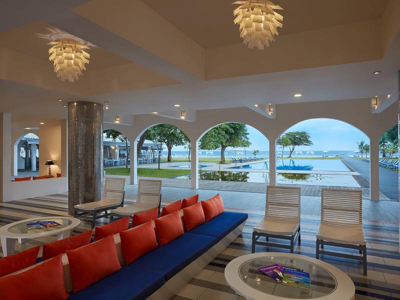 Offener Loungeraum mit roten Kissen, hellen Stühlen und Blick auf Pool und Meer mit Palmen.