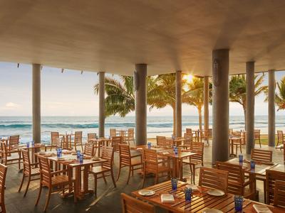 Offenes Strandrestaurant mit Holzstühlen und Tischen, Blick auf Palmen und Sonnenuntergang über dem Meer.