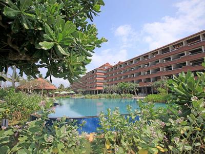 Großer Pool vor mehrstöckigem Hotelgebäude, umgeben von üppigem Grün und blauem Himmel.