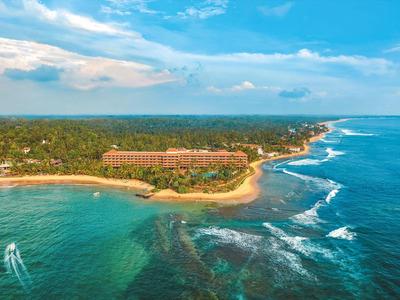 Luftaufnahme eines Küstenresorts mit Sandstrand, Palmen und blauem Meer unter leicht bewölktem Himmel.