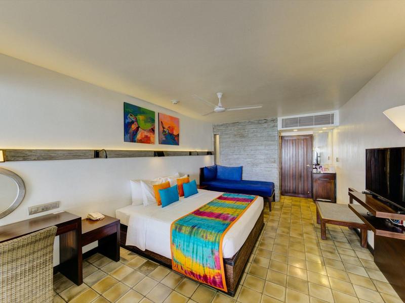 Hotelzimmer mit Doppelbett, bunten Bettakzenten, Sofa, Flachbildfernseher und indirekter Beleuchtung.