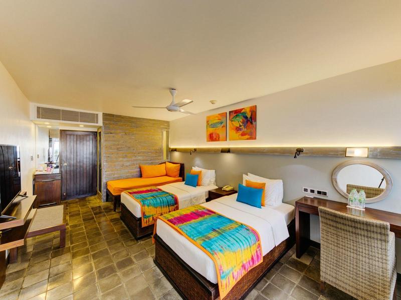 Hell eingerichtetes Hotelzimmer mit zwei Betten, bunter Bettwäsche, Schreibtisch und Fernseher.