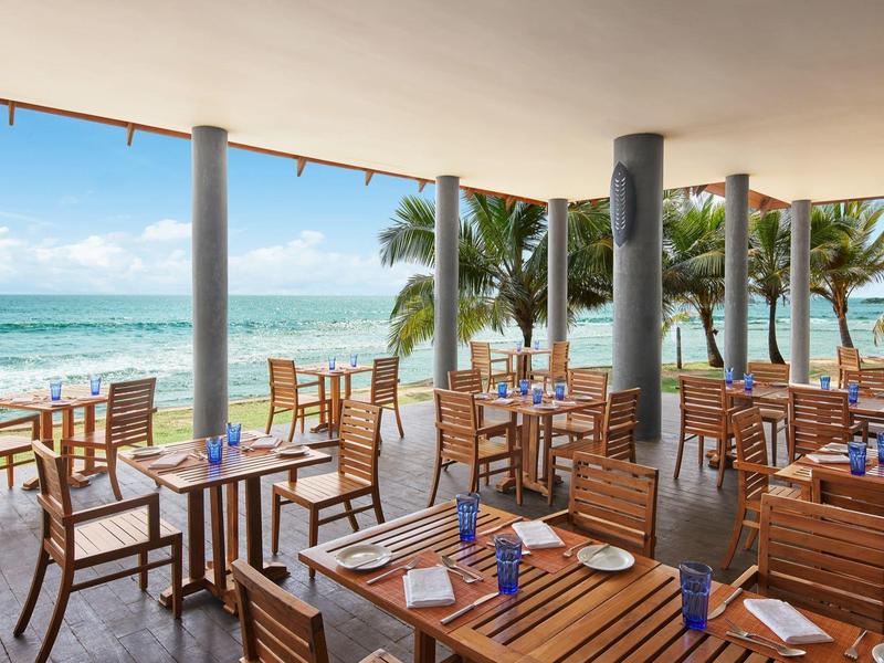 Offenes Restaurant mit Holztischen und Blick auf Meer, blauer Himmel und Palmen am Strand.