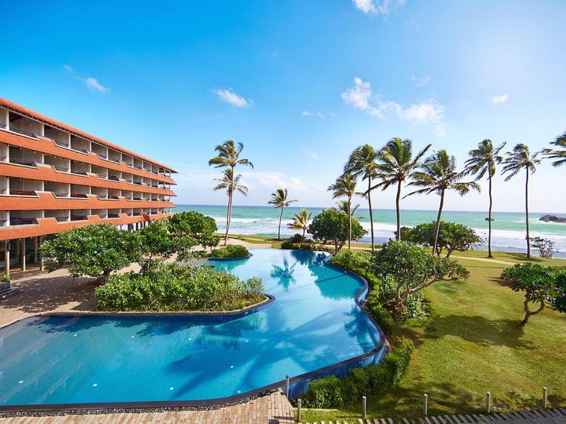 Hotel mit großem Pool, Palmen und Blick auf das blaue Meer unter klarem Himmel.
