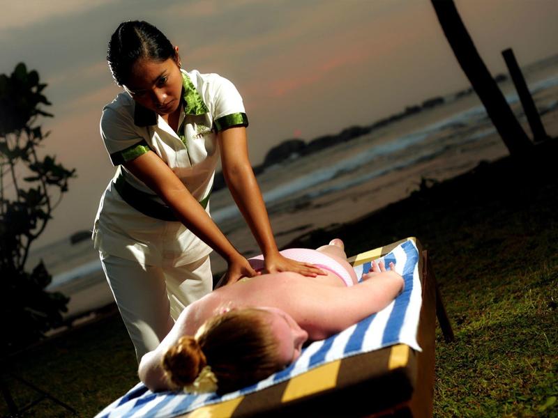 Frau massiert Rücke am Strand bei Sonnenuntergang, ruhige Atmosphäre, gestreifte Liegefläche.