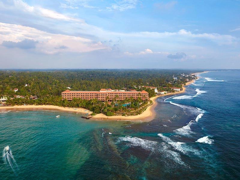 Küste mit Sandstrand, grün bewaldetem Land und einem großen Hotelkomplex unter blauem Himmel.