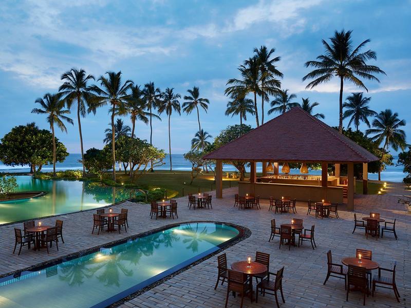 Tropischer Außenbereich mit Pool, Holzmöbeln und Palmen bei Sonnenuntergang am Meer.