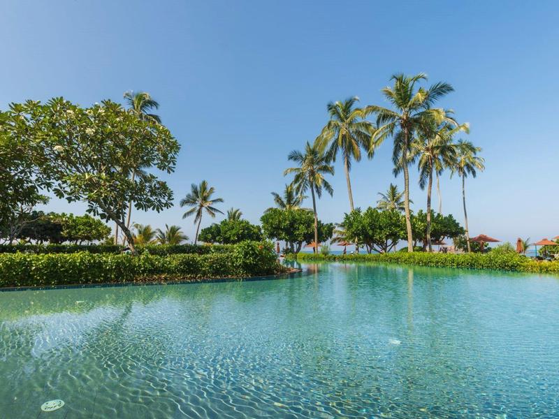 Großer, runder Pool mit blauer Wasserfläche, umgeben von grünen Pflanzen und Palmen unter blauem Himmel.