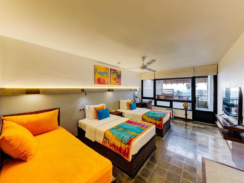 Modernes Hotelzimmer mit drei Betten, bunten Decken, großem Fenster und Flachbildfernseher an der Wand.