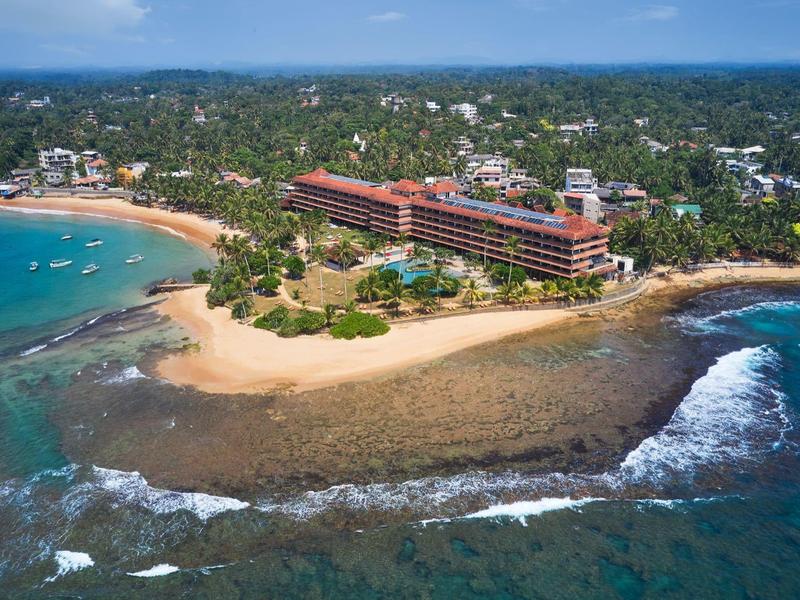 Luftaufnahme eines Hotels am Sandstrand, umgeben von türkisblauem Meer und grüner Vegetation.
