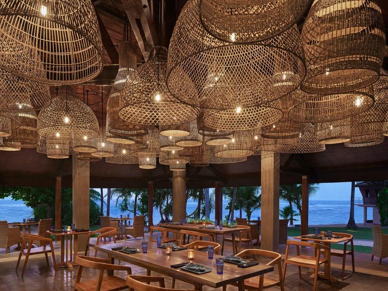 Offener Restaurantbereich mit hölzernen Tischen, Stühlen und großen geflochtenen Lampenschirmen.