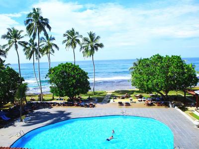 Gran piscina de hotel con palmeras y vista a la playa y el océano al fondo.
