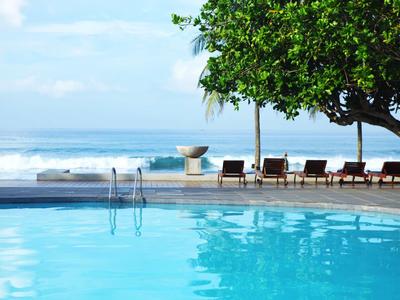 Piscina azul junto a la playa con tumbonas y árbol bajo cielo azul.