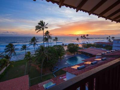 Vista desde un balcón con palmeras, piscina y océano al atardecer en un complejo hotelero.