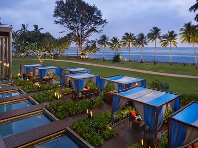 Vista de un paseo frente a la playa con camas cabana azules y palmeras al atardecer.