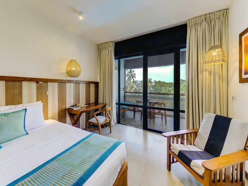 Jasny pokój hotelowy z łóżkiem podwójnym, biurkiem, fotelem i balkonem z widokiem na las.