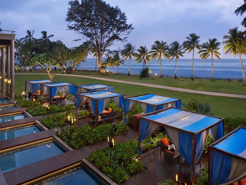 Ogród blisko plaży z niebieskimi zadaszonymi pawilonami i palmami o zmierzchu.