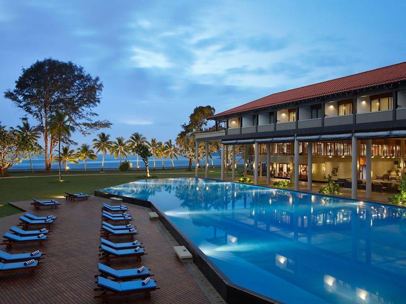 Wieczorny widok hotelu z oświetlonym basenem i leżakami przy wodzie.