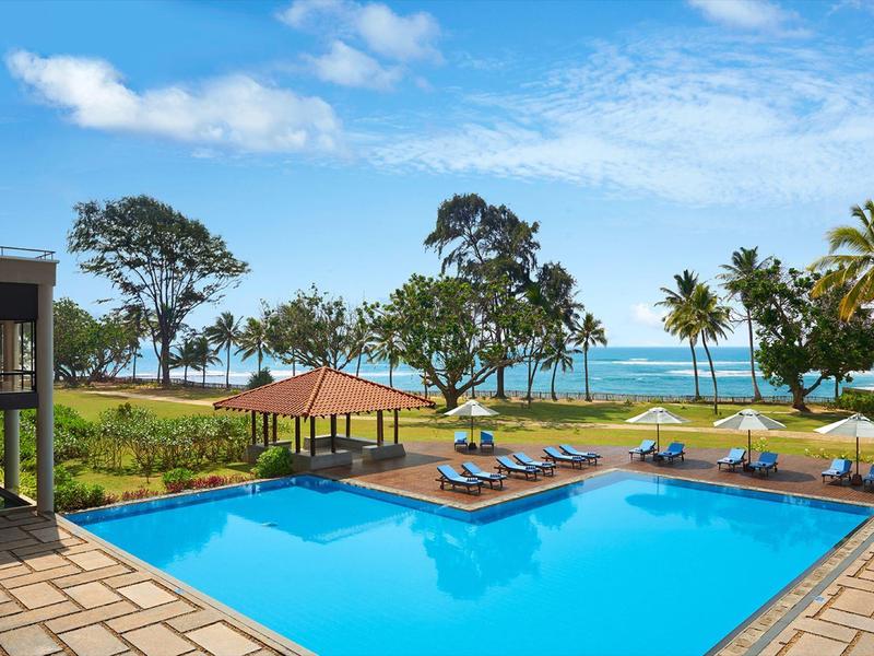 Duży basen z leżakami, otoczony palmami z widokiem na morze i czyste niebo.