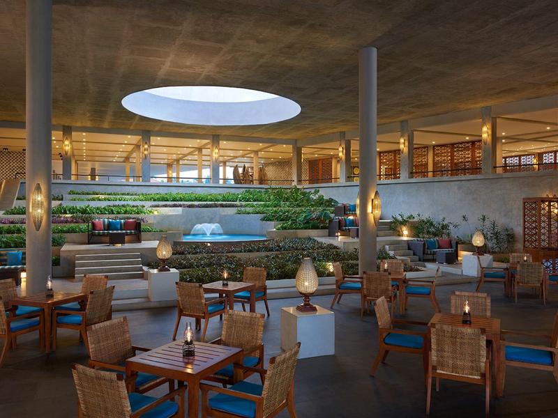 Duża hotelowa lobby z okrągłymi stolikami, krzesłami i zieloną przestrzenią pod okrągłym świetlikiem.