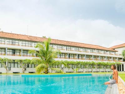 Vrouw zit aan de rand van het zwembad bij een groot hotel met groen planten en palmbomen.