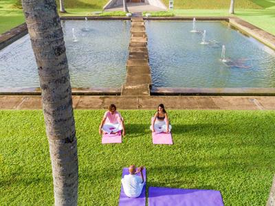 Twee vrouwen die yoga doen op gras met uitzicht op een lang zwembad met fonteinen.