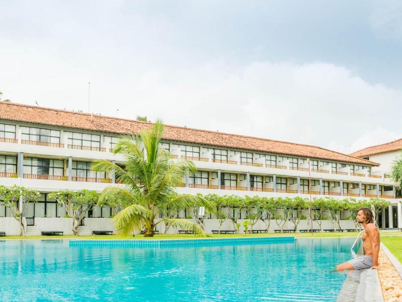 Vrouw zit aan de rand van het zwembad bij een groot hotel met groen planten en palmbomen.
