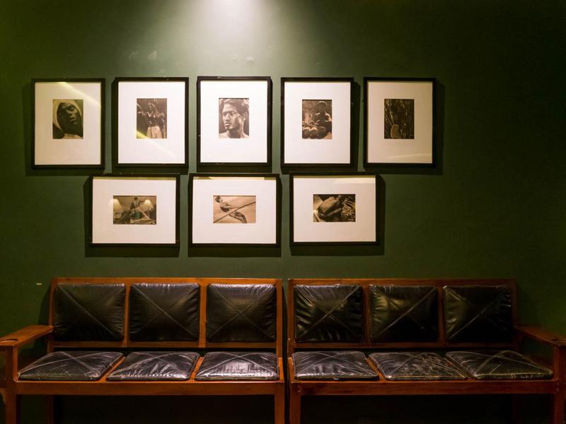 Donkere wachtruimte met leren banken en negen ingelijste foto’s aan een groene muur.