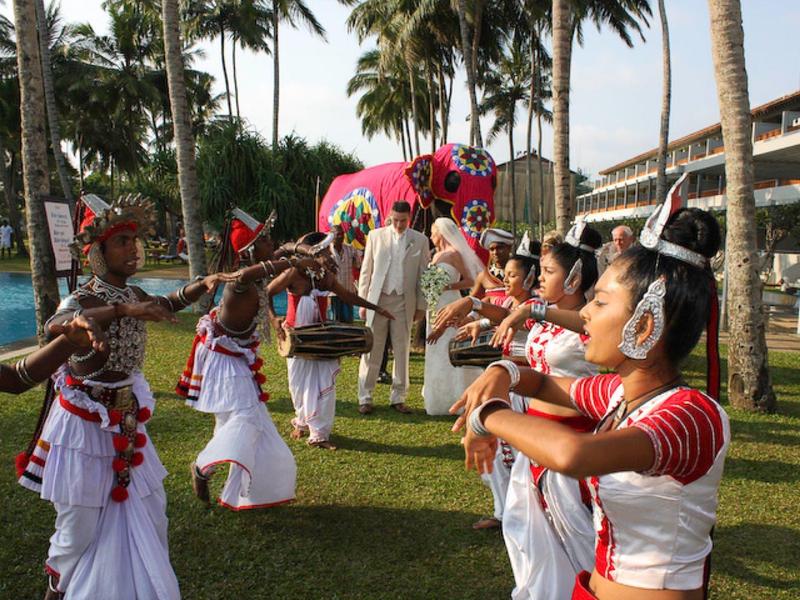 Traditionele dansers in wit-rode kostuums voeren uit voor palmbomen en hotelgebouwen.