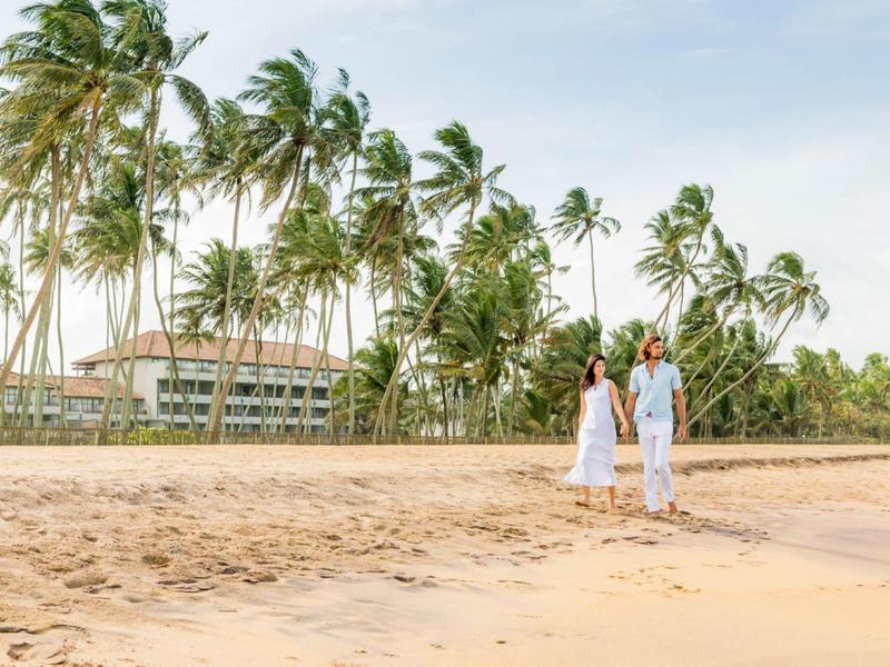 Een stel loopt over een strand met palmbomen en een hotelgebouw op de achtergrond.