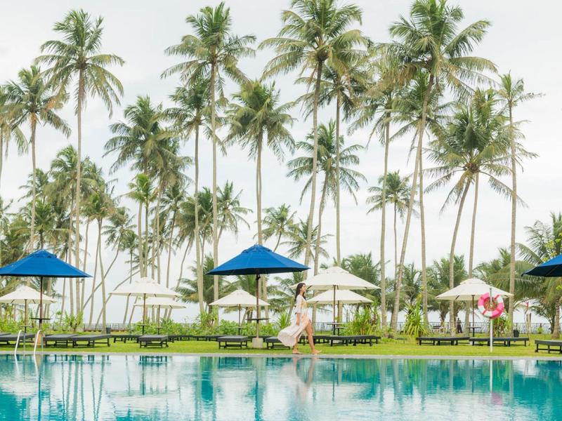 Zwembad met blauwe parasols onder hoge palmbomen bij bewolkte lucht.