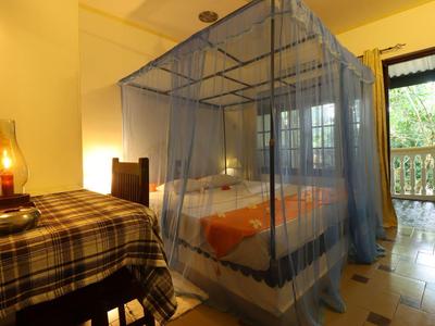 Schlafzimmer mit Himmelbett und blauem Moskitonetz, Holztisch mit kariertem Tuch, offener Balkon.