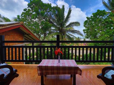 Balkon mit Holztisch, kariertem Tuch und zwei Stühlen, umgeben von grünen Palmen unter blauem Himmel.