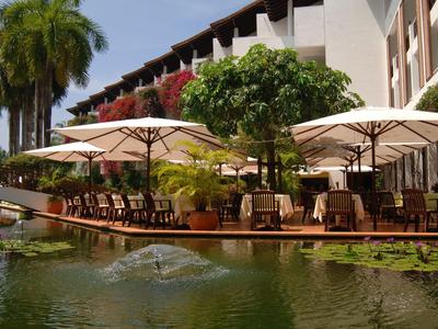 Hotelová terasa s stolky a slunečníky u vody s tropickými rostlinami.