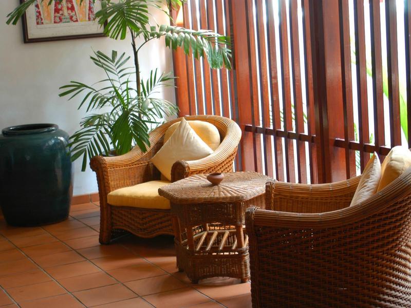 Cozy seating area with rattan chairs, cushions, table, and plants in front of wooden blinds.